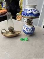 Photo showing two vintage small oil lamps on a surface outdoors: one is a Japanese ceramic oil lamp with blue and white decoration, the other a small brass finger lamp with glass chimney.