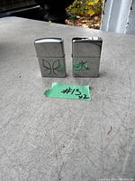 Two Zippo metal lighters placed on a surface showing engraved designs on the front. One lighter has a butterfly or floral pattern while the other shows a scratched letter.