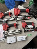 Four Husky pneumatic brad nailers in red and black with metal magazines, two boxes of finishing nails, and a plastic case on outdoor table.
