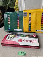 Photo showing three sets of oil paint tubes in original packaging: two sets from Winsor & Newton in green and yellow boxes, and one Georgian Oil Colour set in a red box with tubes and painting knife.