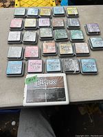 28 Distress Oxide ink pads arranged on table showing a variety of colors, with the distinctive metal casing and color labels, including pink, blue, green, yellow, purple, and grey shades.