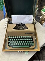 Beige Underwood typewriter with green keys, paper loaded, shown inside wooden carrying case with dark green fabric lining.