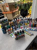 Overview of the full lot showing multiple acrylic paint bottles and tubes on a table outdoors with some stacked boxes in the background.