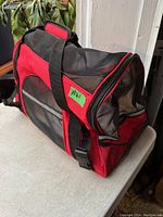 Side view of red and black breathable pet carrier with black mesh panels showing one zipper opening and carrying strap with padded handle.