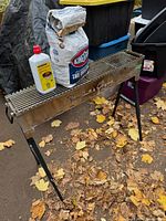 Full view of stainless steel folding BBQ with three black adjustable legs set up outdoors, with bag of charcoal and starter fluid placed on grill surface.