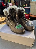 Pair of ladies winter boots displayed on white shoebox outdoors. Taupe boots with snake print overlays and faux shearling lining showing. Lace-up boots with metal eyelets and hooks.