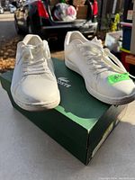 Pair of white Lacoste sneakers on top of green Lacoste box, showing front and top profile of shoes. White mesh and synthetic uppers with white laces.