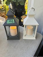 Photo of two battery operated lanterns, one black and one white, both with flickering LED candles inside. Both lanterns are about 9.5 inches tall with handles on top.