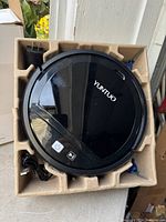 Top view of black Yuntuo smart vacuum robot inside an open box with power adapter and cable.