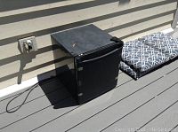 Black compact bar fridge placed on outdoor wooden deck next to cushions.