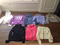 Eight girls sweatshirts, sweaters and down vest laid out on wooden floor showing variety and color.