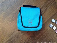 Closed blue Nintendo DS carrying case with black trim and shoulder strap on wooden floor.