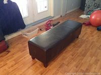 Full view of a long black faux leather bench on wooden flooring with short legs and some surrounding household items.