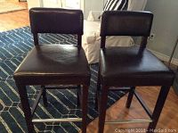 Front view of two brown faux leather bar stools with wooden legs and metal footrest, placed on patterned carpet.