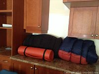 Photo showing navy blue and red sleeping bag, orange rolled tent pad secured with black straps, and large black cushion on kitchen counter.