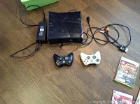 Photo showing Xbox 360 console, power supply, AV cable, and two wireless controllers (black and white).