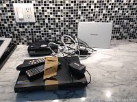 Electronics including black LG DVD player, white Netgear modem, black clock radio, two remote controls and power adapters on counter with tiled backsplash.