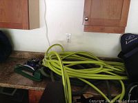 Photo showing two long garden hoses; one light green and brand new, the other dark green and used. Also present are two metal and plastic oscillating sprinklers on a countertop under wooden cabinets.