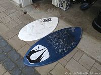Two skim boards on a paved area, a white board and a worn blue board with taped separation at front.