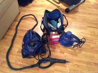 Blue electric steamer with attached hose, power cords, blue car polisher, two pink containers of car wax, and a blue carry bag on a wood floor.