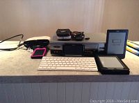 Photo showing front-line electronics including Kindle e-readers, Apple keyboard, VCR, camera, and media player on a countertop.