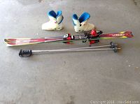 Photo shows full ski set including skis with bindings, boots, and ski poles on concrete floor.