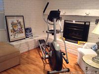 Wide side-angle view of the elliptical trainer showing the full frame, rails, and handlebars in a room setting by a fireplace.