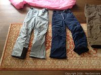 Five pairs of ski pants laid on floor, showing three pairs: light gray, navy blue, and brown pants