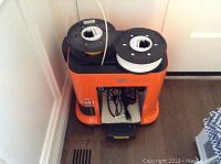 Front-angle view of orange 3D printer showing top spool holders with yellow and white spools