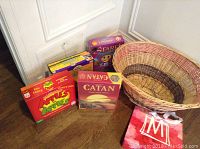 Photo showing five boxed board games including Catan, Battle Ship, Cranium, Safari, a wicker basket, and a pink shopping tote bag with white 'M' on it.
