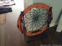 Photo of folded orange trampoline chair with black bungee netting on a gray metal frame, showing compact storage state.
