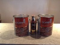 Two full cans of Olympic Deck, Fence & Siding stain and sealant in semi-transparent finish with one can of Krylon Maxx metallic spray paint