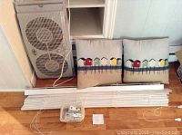 Photo showing Hampton Bay box fan on its side, two cushions with house pattern covers, and white horizontal blinds with cords and accessories on a wooden floor.