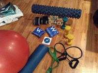 Photo of all exercise equipment laid on wooden floor showing stability ball, foam roller, dumbbells, resistance bands, wrist supports, yoga mats, and DVD/manual packs.
