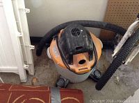 Photo of used orange and gray shop vac with attached black hose coiled on top, showing wear from use and power cord wrapped around top.