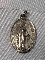 Front view of the oval sterling silver pendant charm showing a raised figure of the Virgin Mary with inscribed border text