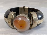 Front view of the bracelet showing large round polished central stone with orange and white color within a silver-tone setting and the dark wide band.