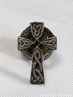 Close-up photo of silver-tone Celtic cross pin showing knotwork design on cross arms and stem against white fabric background.