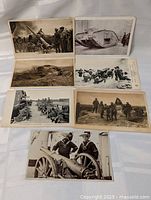 Seven WW1 vintage postcards laid out on a white cloth showing various military scenes and equipment.