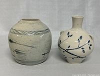 Two pottery vases side by side showing overall shape and design. The larger vase has abstract blue brushline decorations and the smaller vase has a blue branch and leaf pattern with handles.