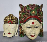 Two vintage hand carved wooden Balinese masks side by side, showing the front detail of the larger mask with intricate colorful headdress and facial features.