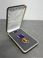Opened display case showing replica Purple Heart medal and matching ribbon bar nestled in grey foam and white satin lining.