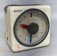 Front angled view showing square clock with cream-colored body, red alarm hand, blue minute hand, yellow second hand, and Sony logo on face.
