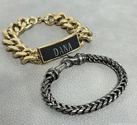 Photo of gold chain bracelet with black 'DANA' nameplate and silver woven chain bracelet placed on gray surface.