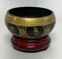 Front view of the singing bowl with detailed gold inscriptions and patterns on a black surface, resting on the wooden stand.
