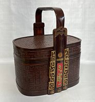 Front angle view of vintage Chinese bamboo lunch box with dark reddish finish, showcasing the curved handle and ornamental brass hardware with red floral enamel panels.