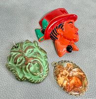 Three vintage jewelry pieces on gray textured surface: red-green painted brooch of man's head, green theatrical face brooch, and oval painted pendant of three women