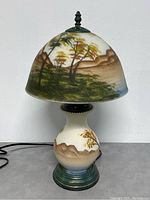 Front view of lamp showing full scenic reverse painted glass shade and base with brown mountains, green/yellow trees, blue water, and green base.
