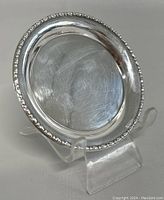 Front angled view of small round sterling silver dish on clear display stand, showing polished surface and beaded rim detail.