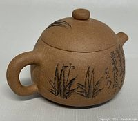 Angled side view of the mini teapot showing handle, botanical etching, and calligraphy script on clay surface.